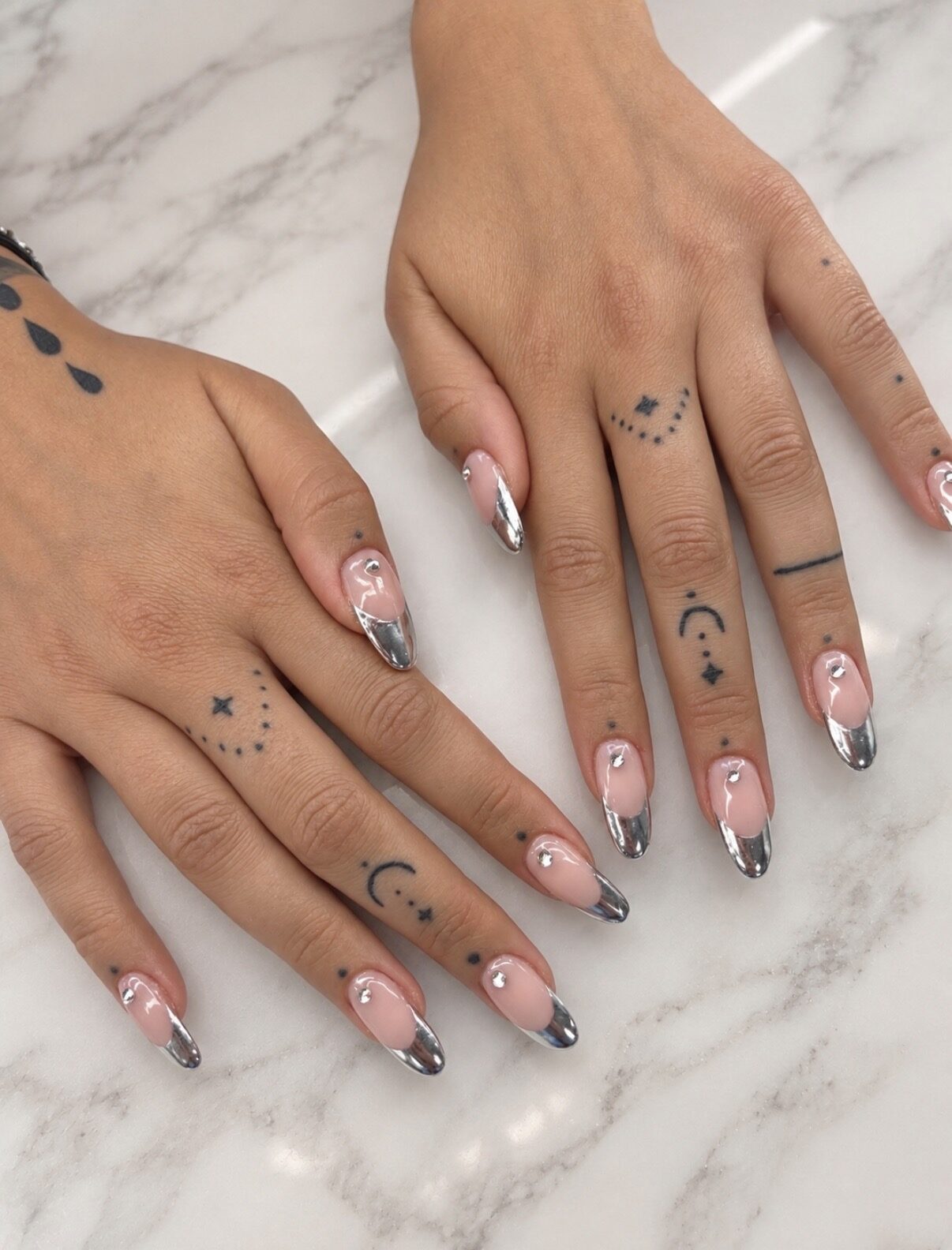Francesa plateada chrome sobre uñas largas, manos tatuadas, sobre mármol.